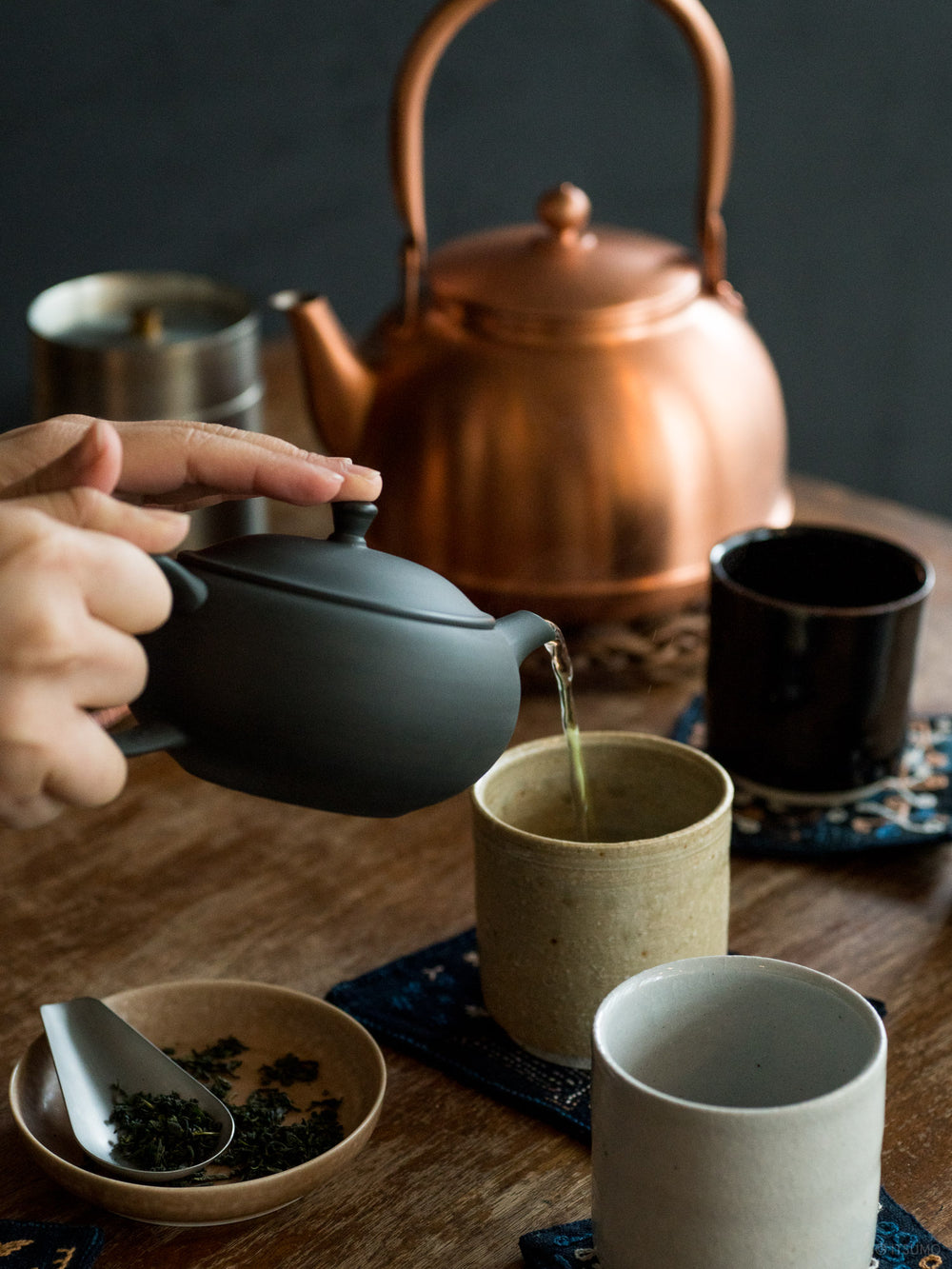 Azmaya tokoname ware black teapot pouring tea into ceramic cups with a copper kettle in the background