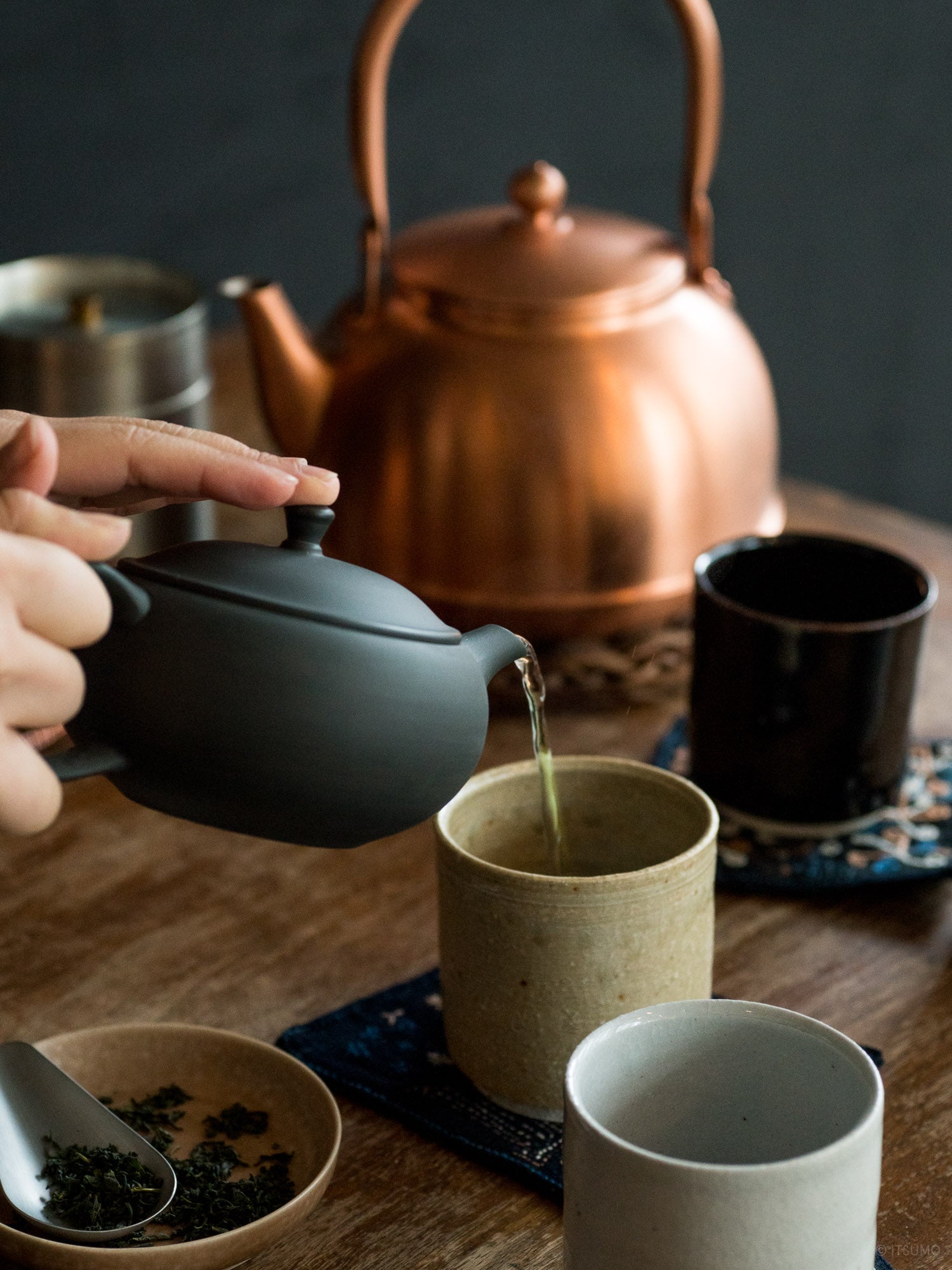 Azmaya black tokoname ware oval teapot pouring tea into a ceramic cup with copper kettle in the background