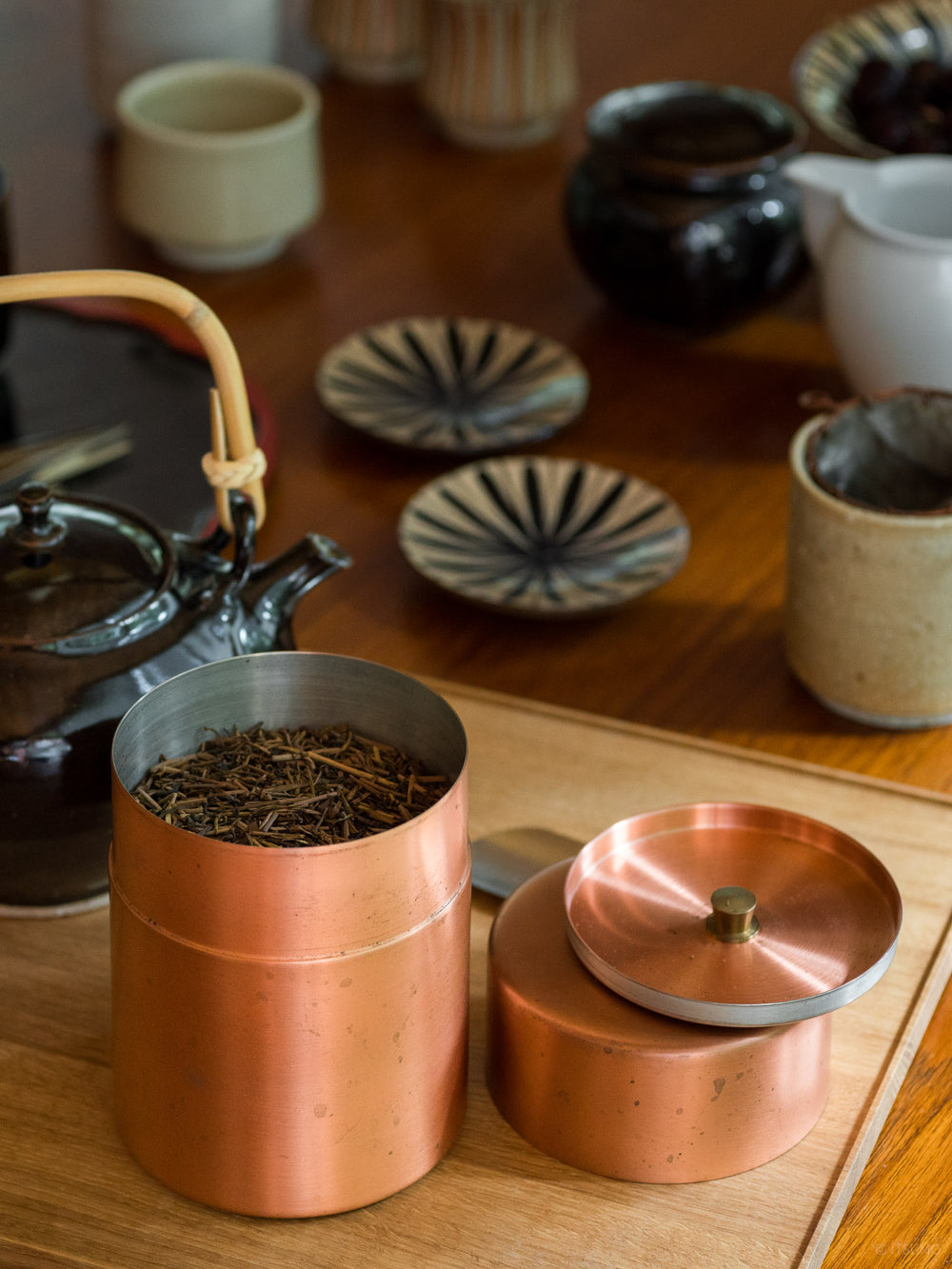 Azmaya copper tea canister filled with tea leaves next to a ceramic kettle and cups