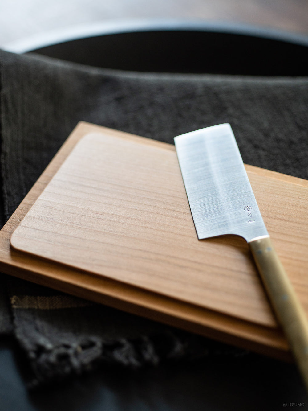 Azmaya stainless steel cheese knife with a brass handle sitting on top of a wooden cutting board