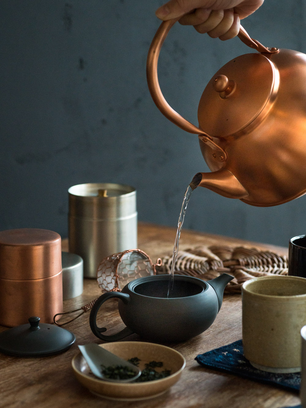 Copper kettle pouring tea into a teapot next to ceramic cups and copper tea canisters