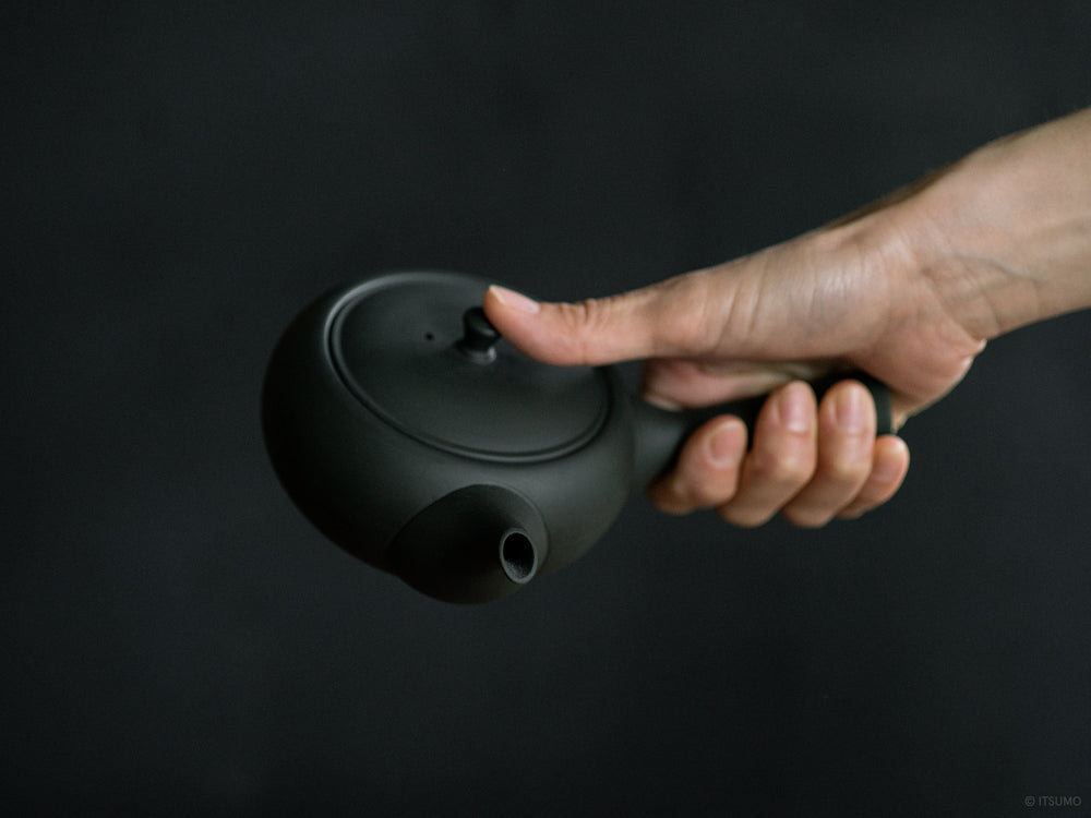 Hand holding Azmaya ceramic oval teapot in unglazed matte black with side handle